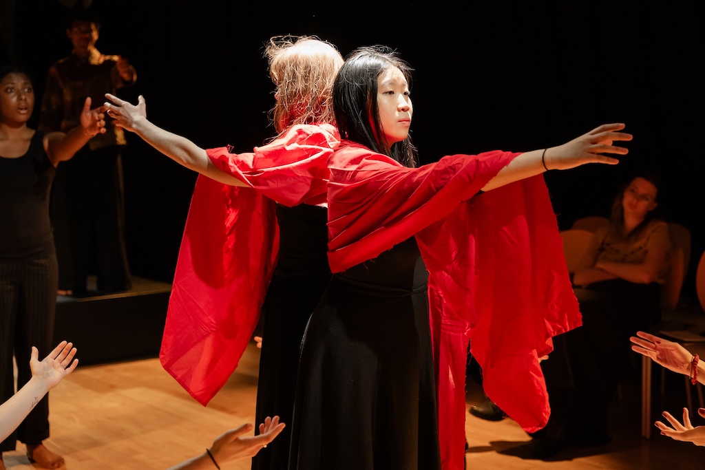 Two actors wearing bright red capes stand with their backs to each other with their arms outstretched in front of them.